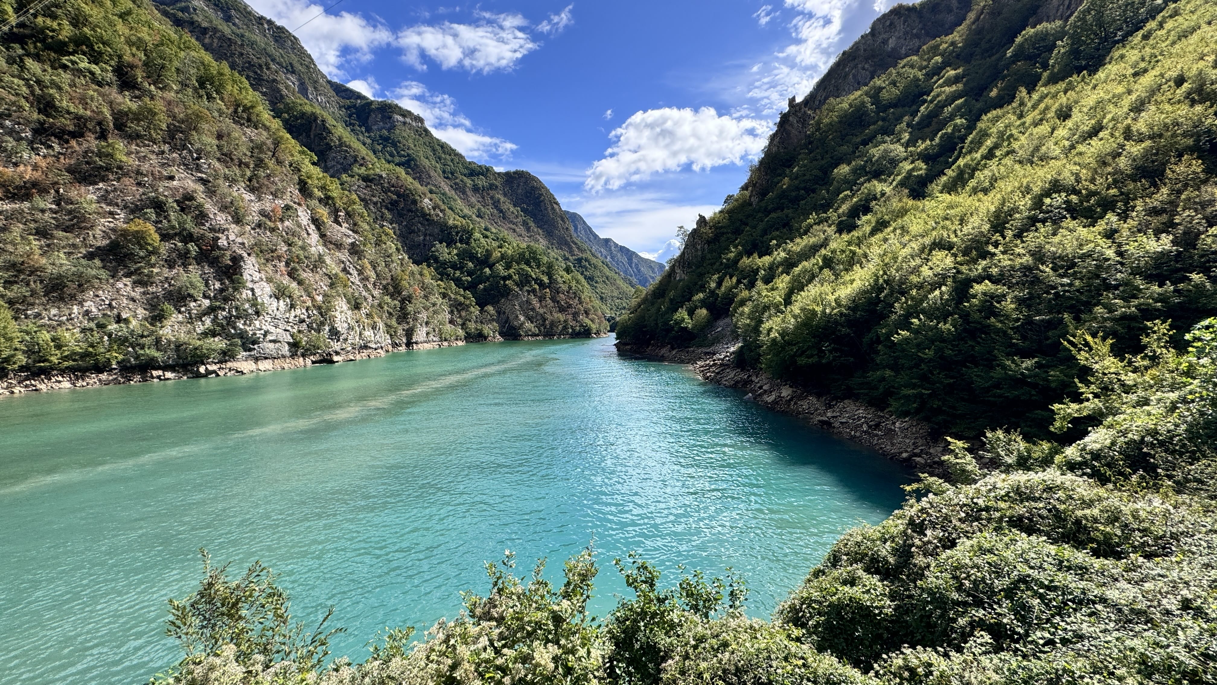 Turquoise waters of Komani Lake surrounded by steep, rocky cliffs and forested mountains under a bright blue sky in northern Albania.