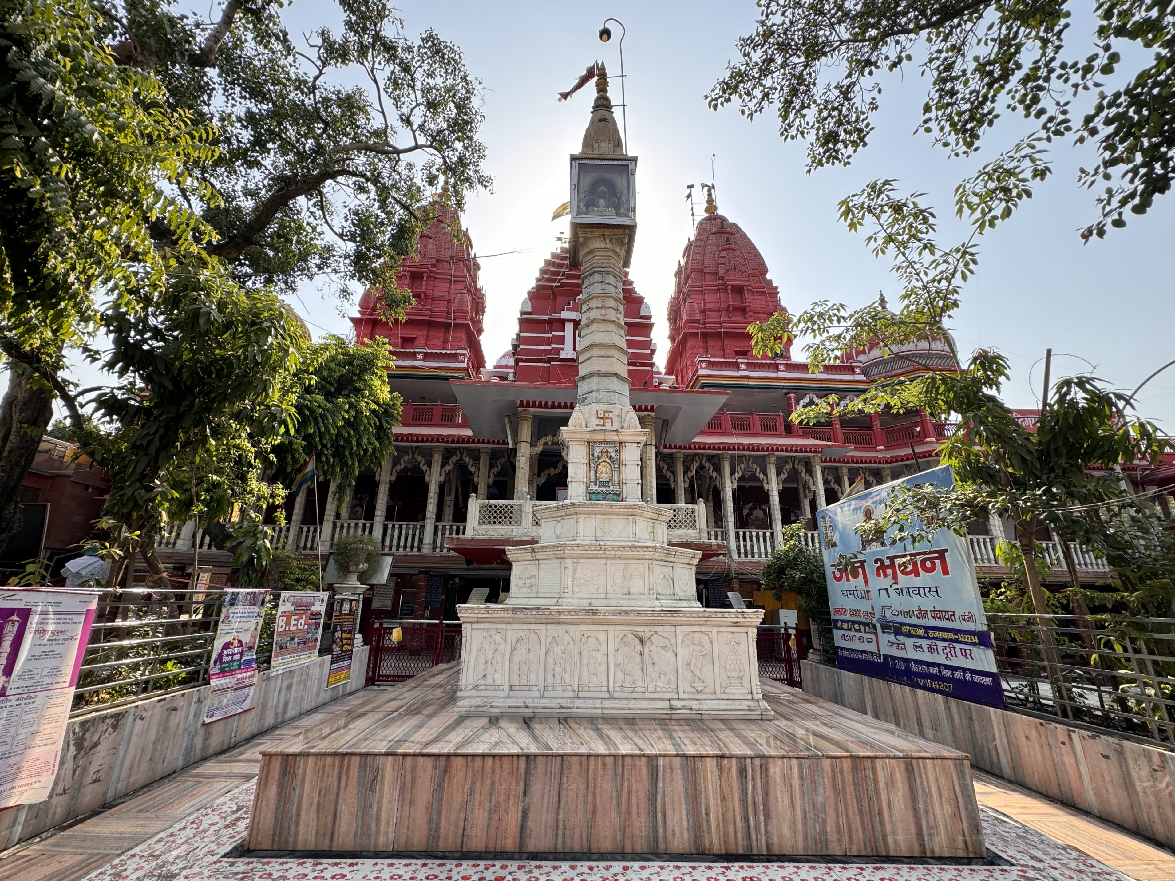 Visitors stand before the marble column and bright red spires of Shri Digambar Jain Lal Mandir, Delhi’s oldest Jain temple, framed by leafy trees and religious posters.