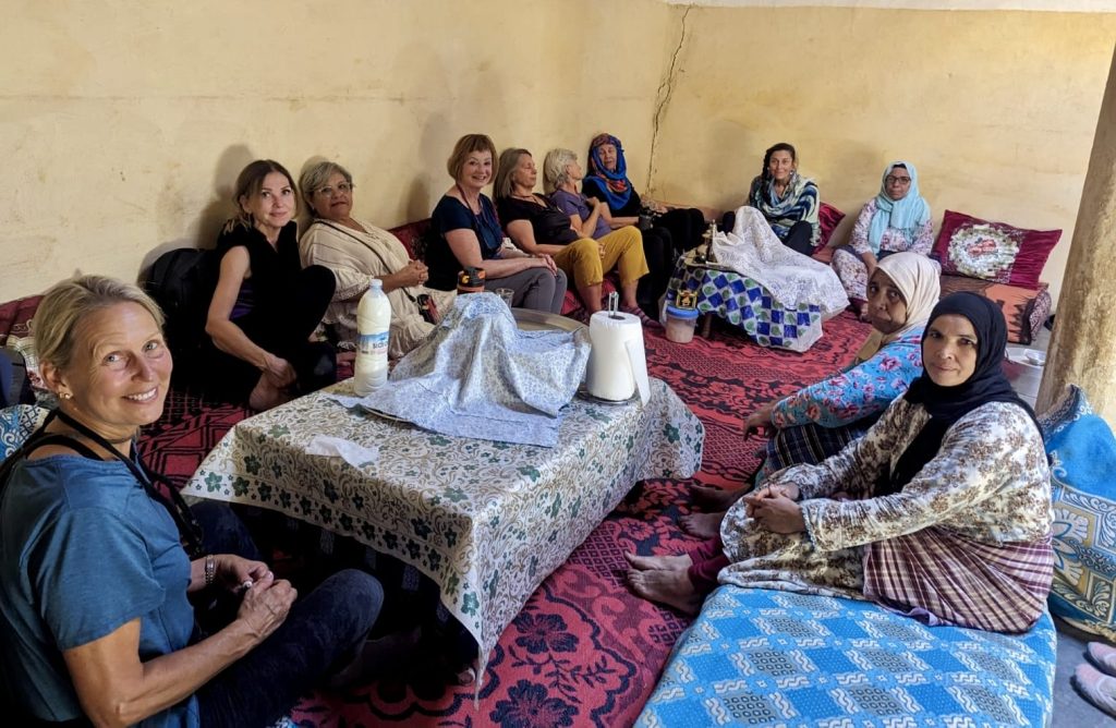 A group of foreign women travelers sit on floor cushions around a low table, sharing space with local Moroccan women from the El Waha cooperative, inside a simple room with pale yellow walls and patterned rugs.