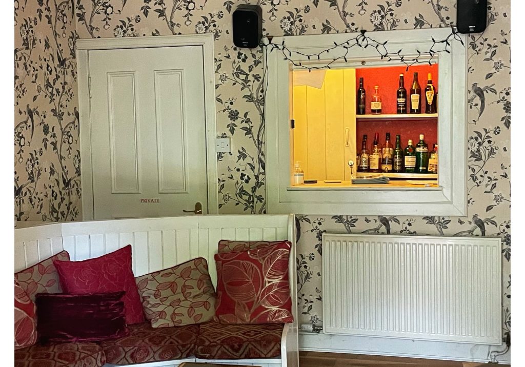 A cozy sitting room with patterned wallpaper and a bar window showcasing bottles of whiskey and spirits.