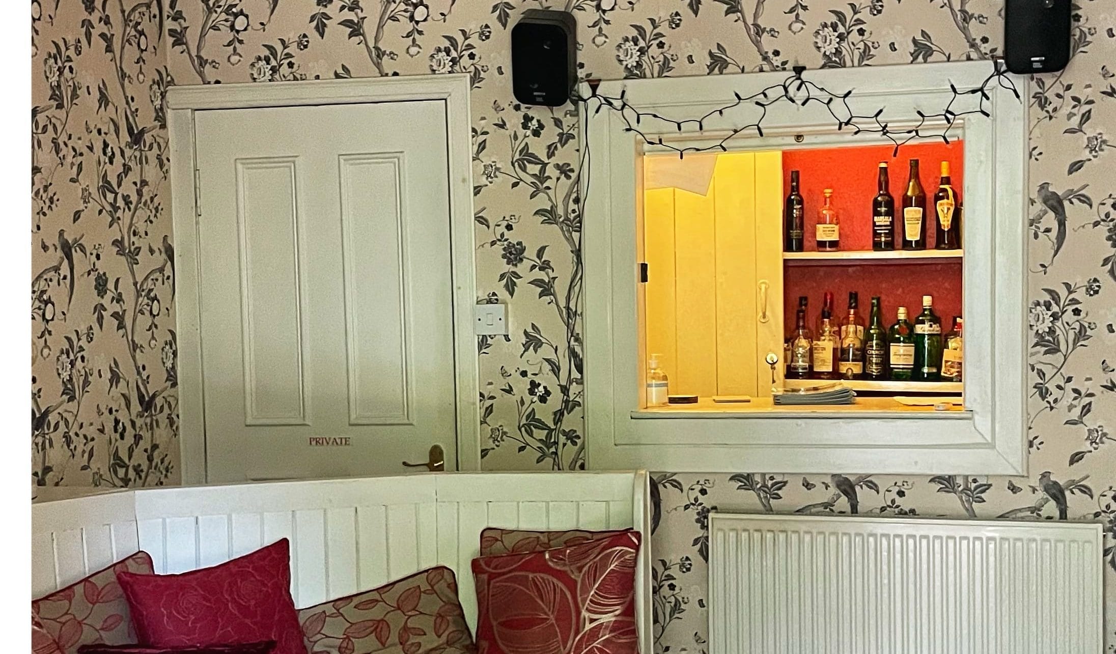 A cozy sitting room with patterned wallpaper and a bar window showcasing bottles of whiskey and spirits.
