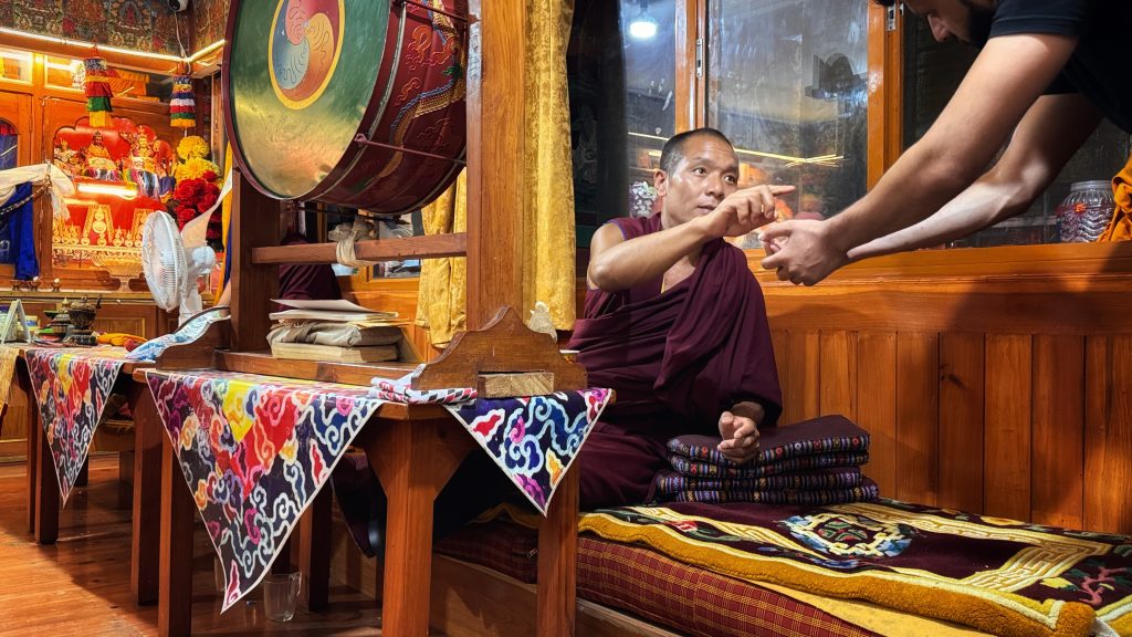 Tibetan monks seated in a prayer room chanting during a puja ceremony, surrounded by ritual objects.