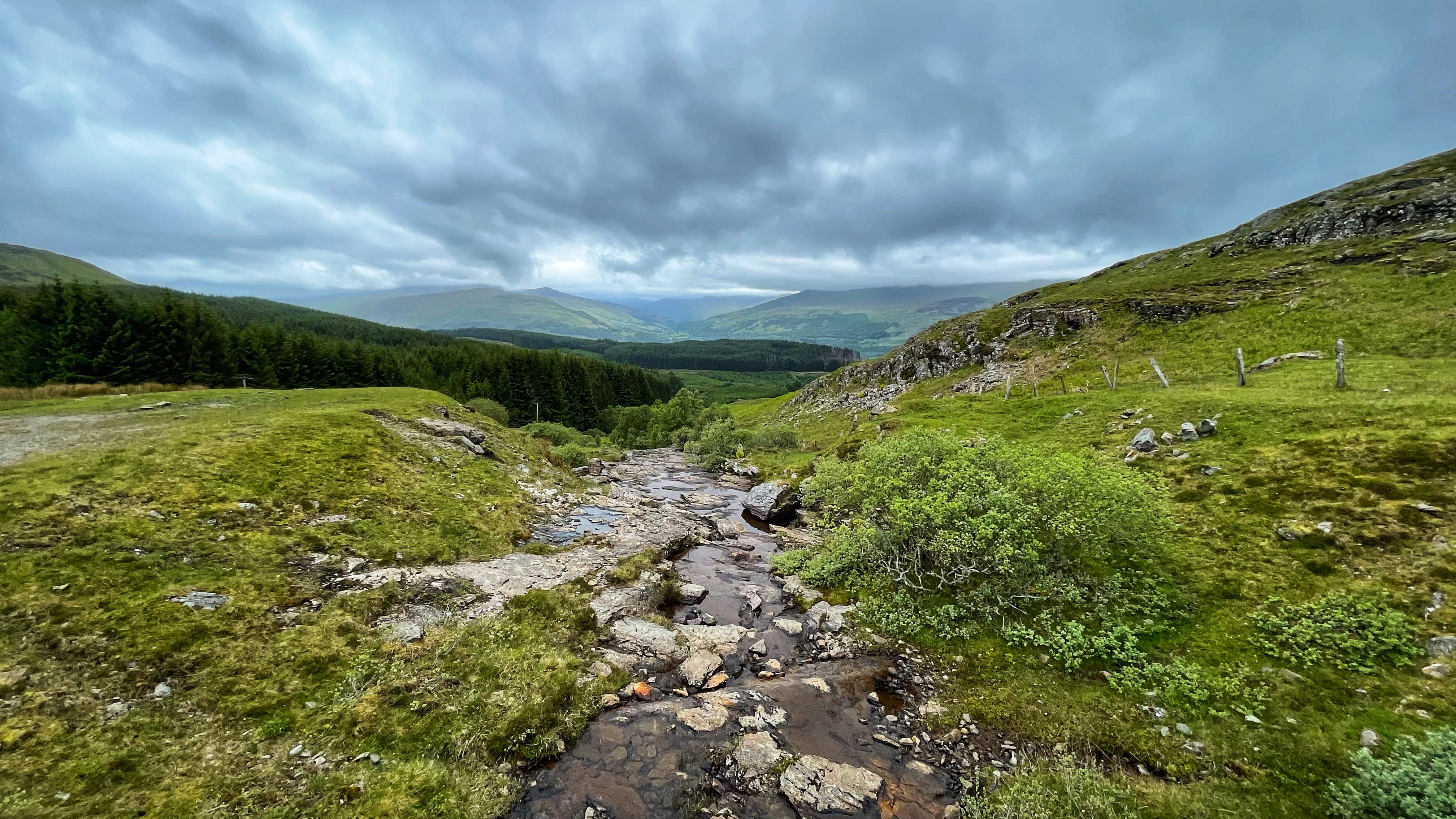A rocky stream flows through a mossy green hillside, with dark pine forests and distant mountains under dramatic, cloud-filled skies in the Scottish Highlands.
