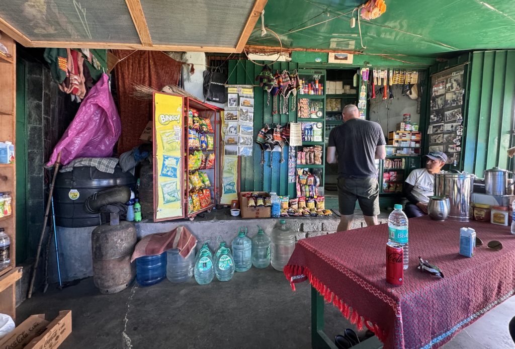 A rustic shop interior with shelves of snacks, water jugs, a red tablecloth-covered table, and a man ordering from the counter.