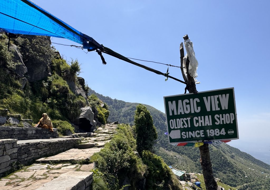 A hiker walks past a sign that reads “Magic View Oldest Chai Shop Since 1984” on a stone path overlooking the mountains near Triund.