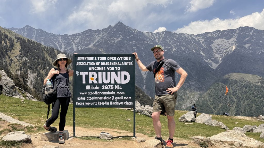 Woman and man stand on either side of the Triund sign at the summit, framed by dramatic mountain peaks and a clear alpine sky.