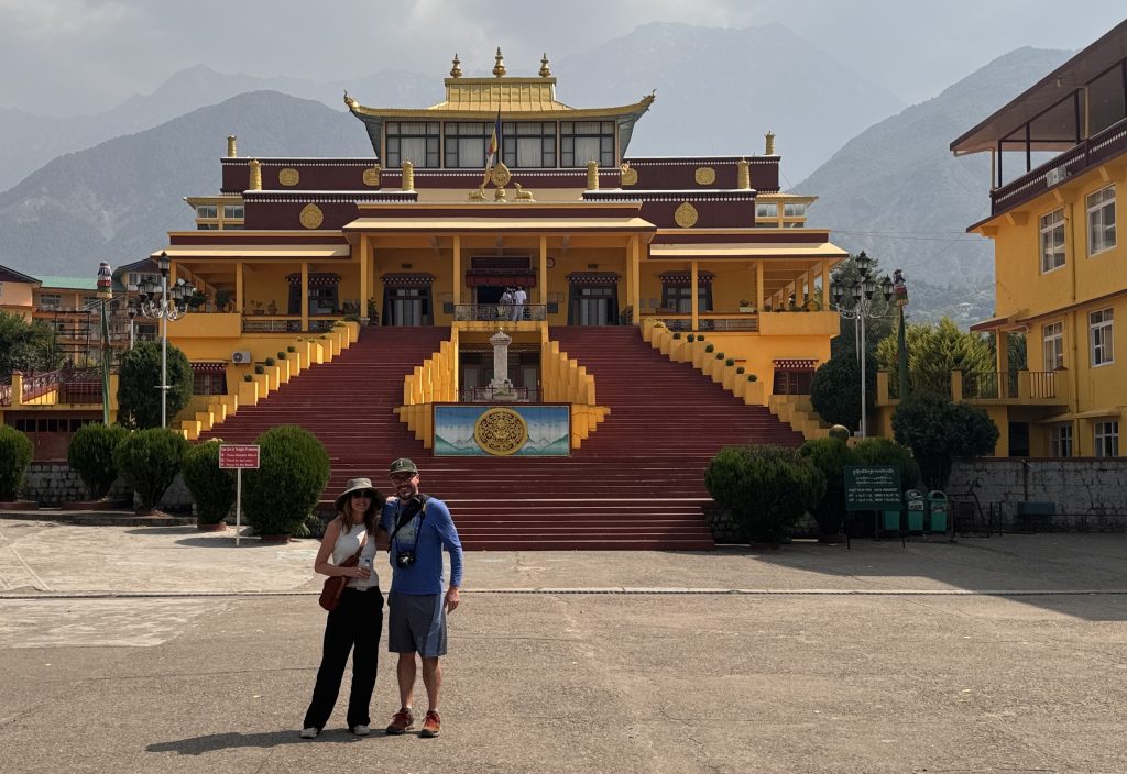 A couple stands in front of the grand entrance to Gyuto Monastery, with red steps, yellow walls, and golden rooftops framed by Himalayan mountains.