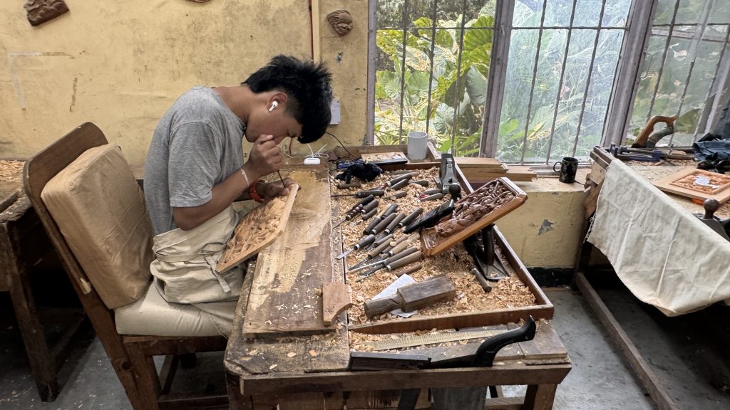 Young Tibetan artisan carving intricate designs into a wooden panel at a tool-covered workbench.