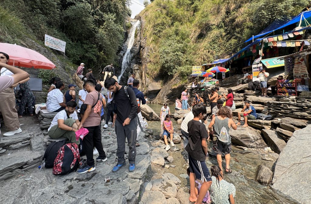 A busy scene at the base of Bhagsu Waterfall, with visitors relaxing on rocks and wading in the stream, surrounded by snack stalls and prayer flags