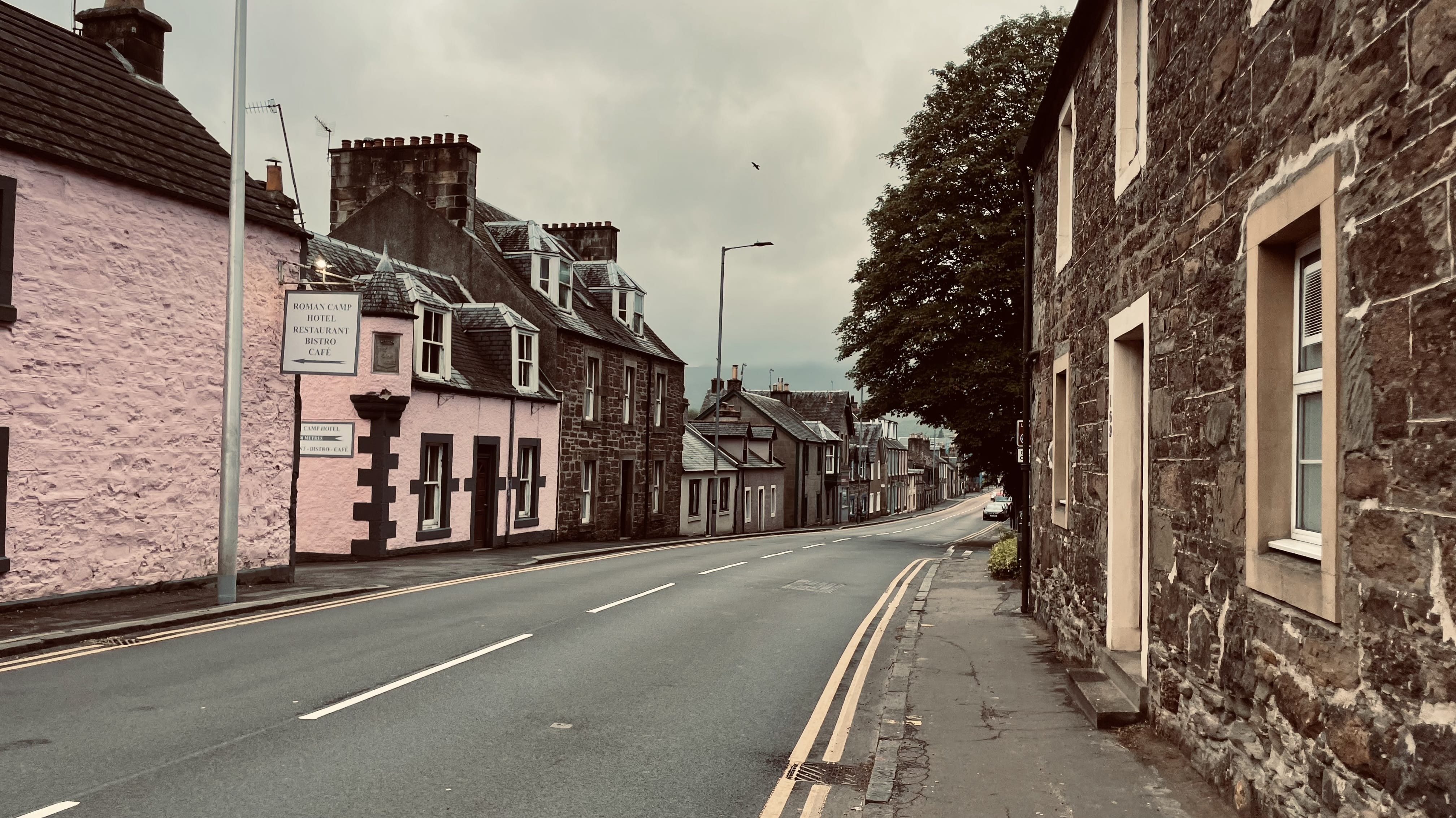 Pastel-colored stone houses, including a pink hotel with a sign for Roman Camp Hotel Restaurant Bistro Café, along a gently curving road in Callander.
