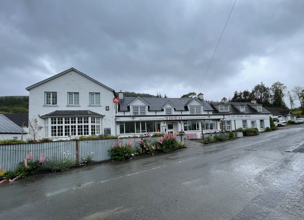 A white roadside inn called Mhor 84 with lupines blooming out front and rain clouds above.