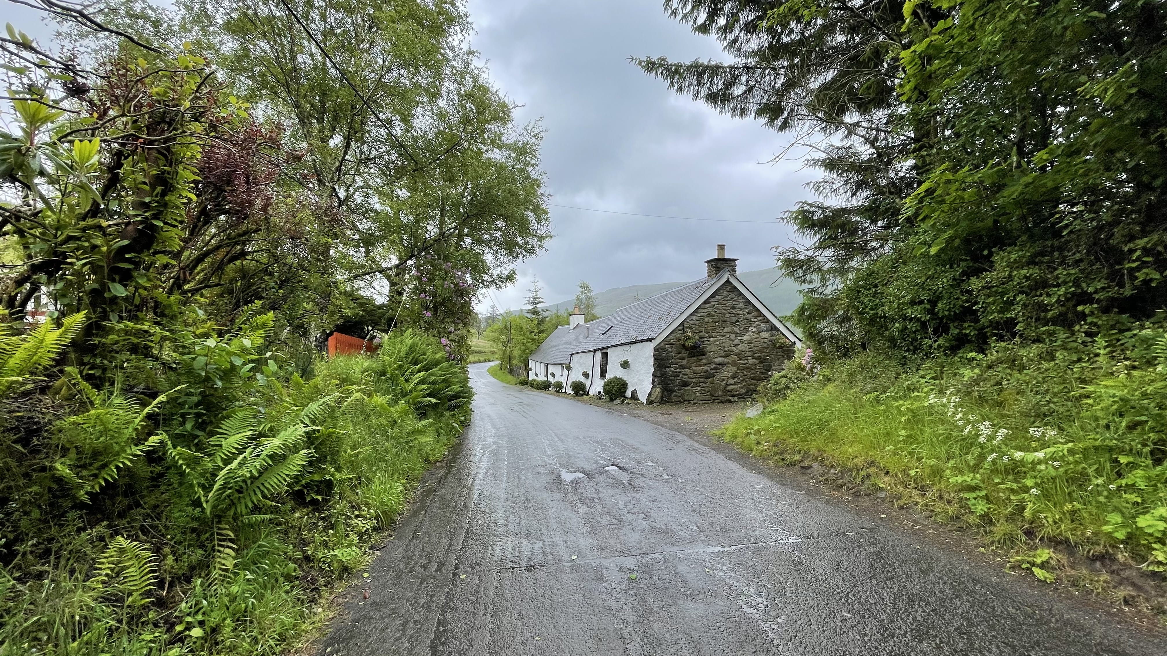 A narrow, rain-slick road curves past a white stone cottage surrounded by greenery on a cloudy day while hiking the Roy Roy Way, Scotland