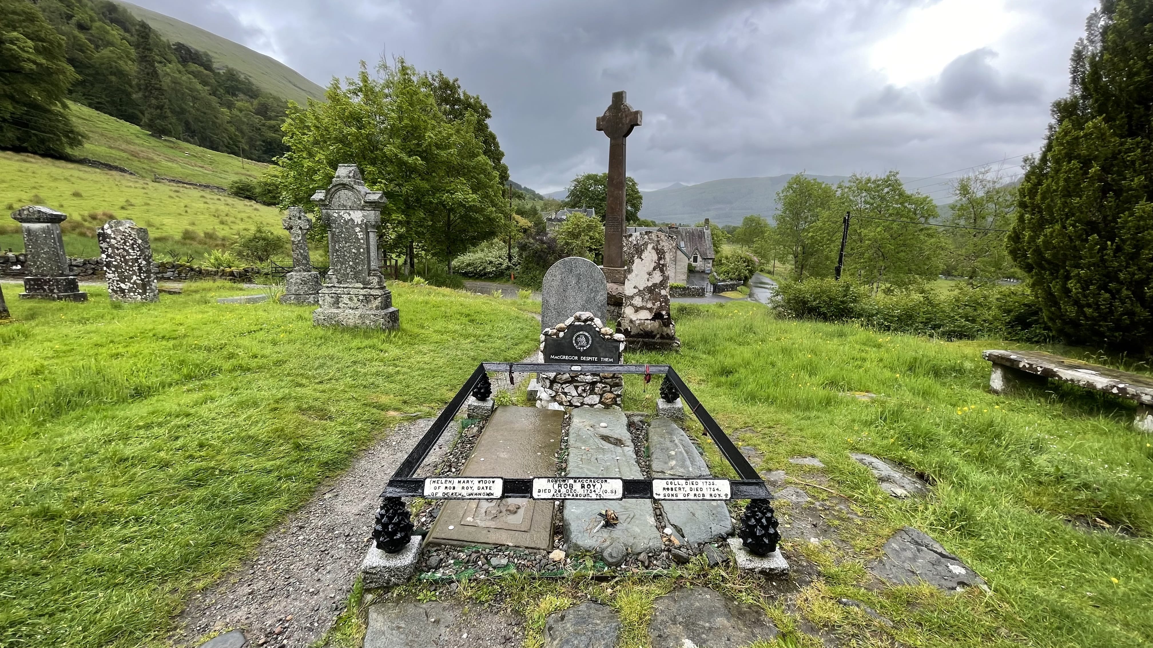 Historic grave marked “MacGregor Despite Them All” surrounded by other weathered headstones and hills in the distance.