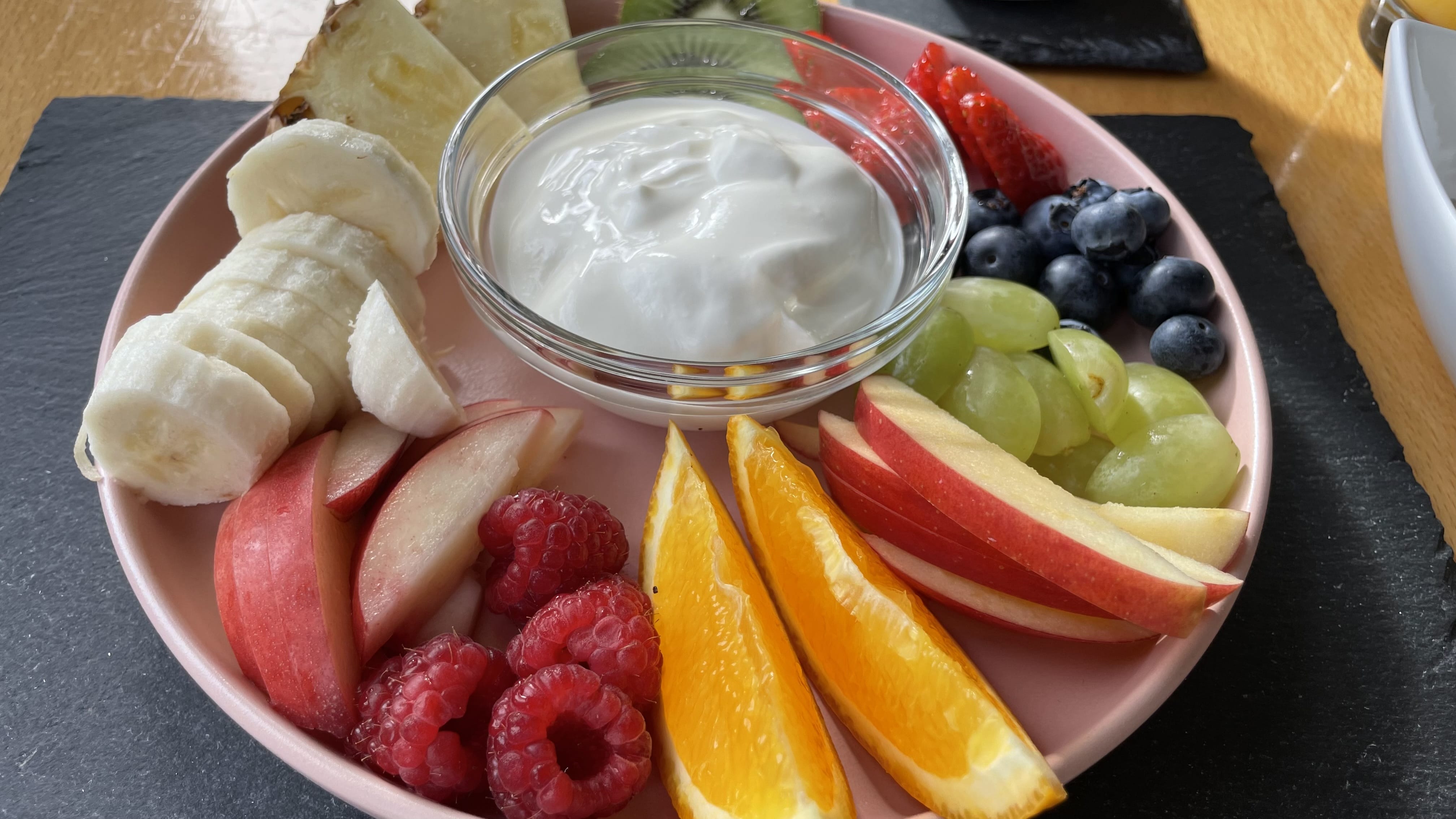 A colorful plate of sliced banana, apple, orange, raspberries, strawberries, kiwi, pineapple, blueberries, and grapes with a bowl of plain yogurt in the center.