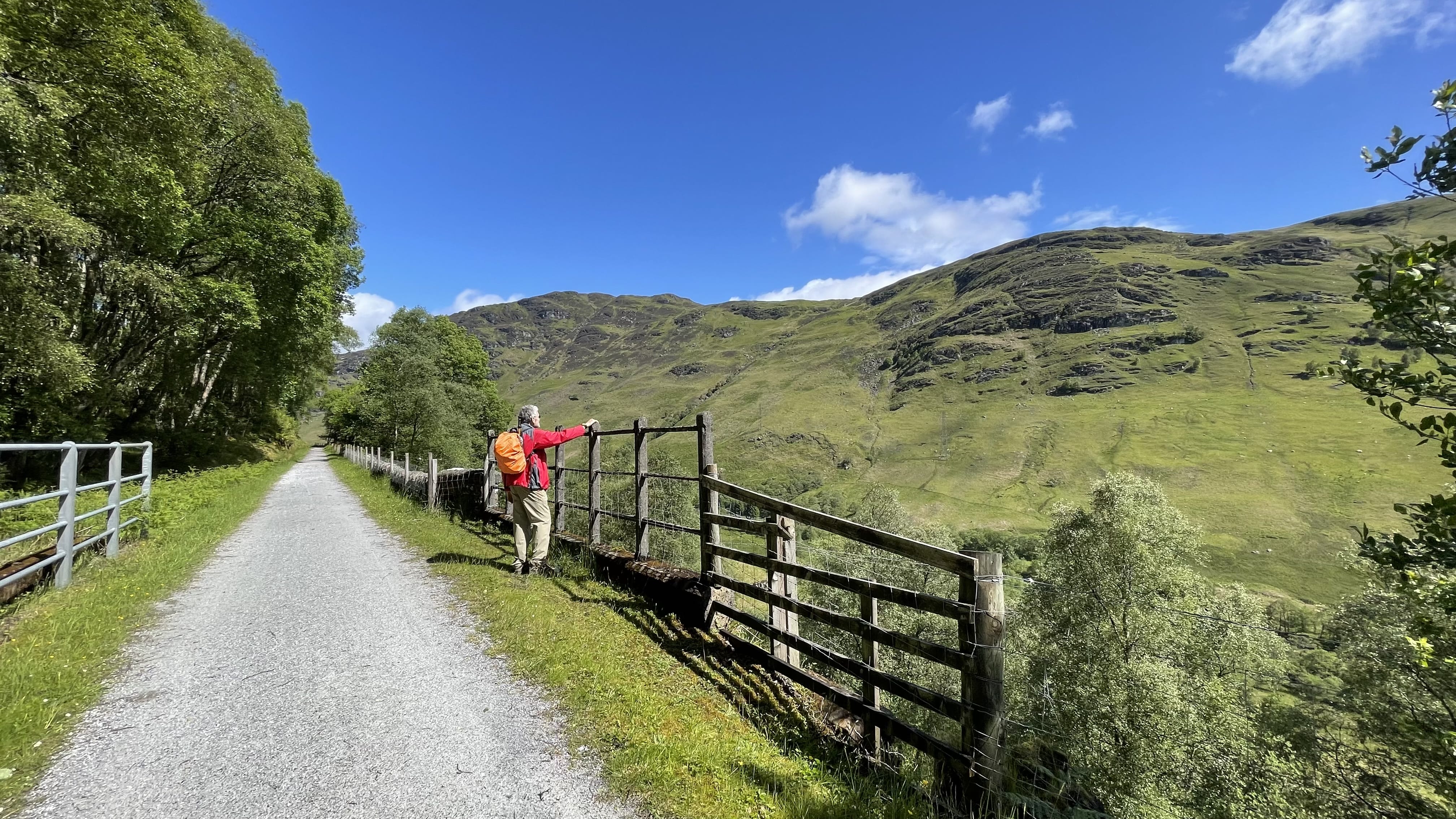 A hiker in a red jacket and orange backpack pauses at a wooden fence along the Glen Ogle trail, looking out across sunlit green hills under a bright blue sky.