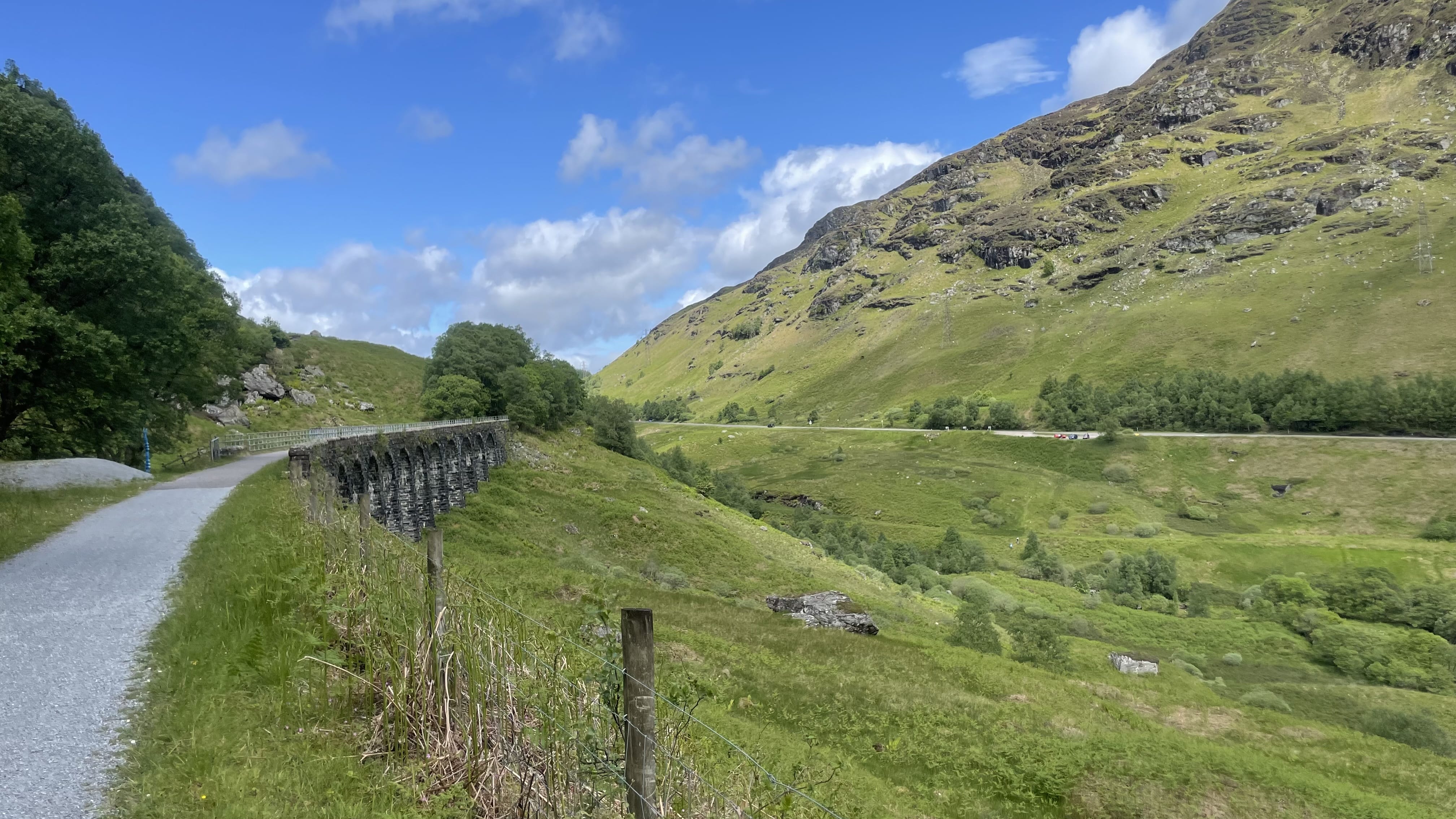 Stone viaduct curving through the lush green valley of Glen Ogle, with a walking path beside it and rolling hills under a blue sky while hiking the Roy Roy Way, Scotland