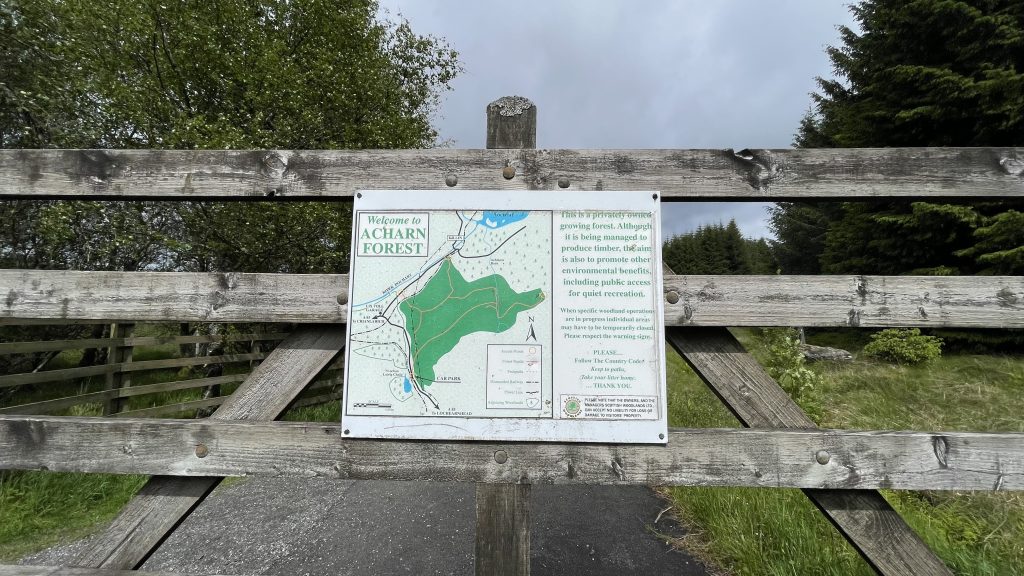 Signboard at the entrance to Acharn Forest, mounted on a wooden gate, showing a trail map and visitor information surrounded by trees.