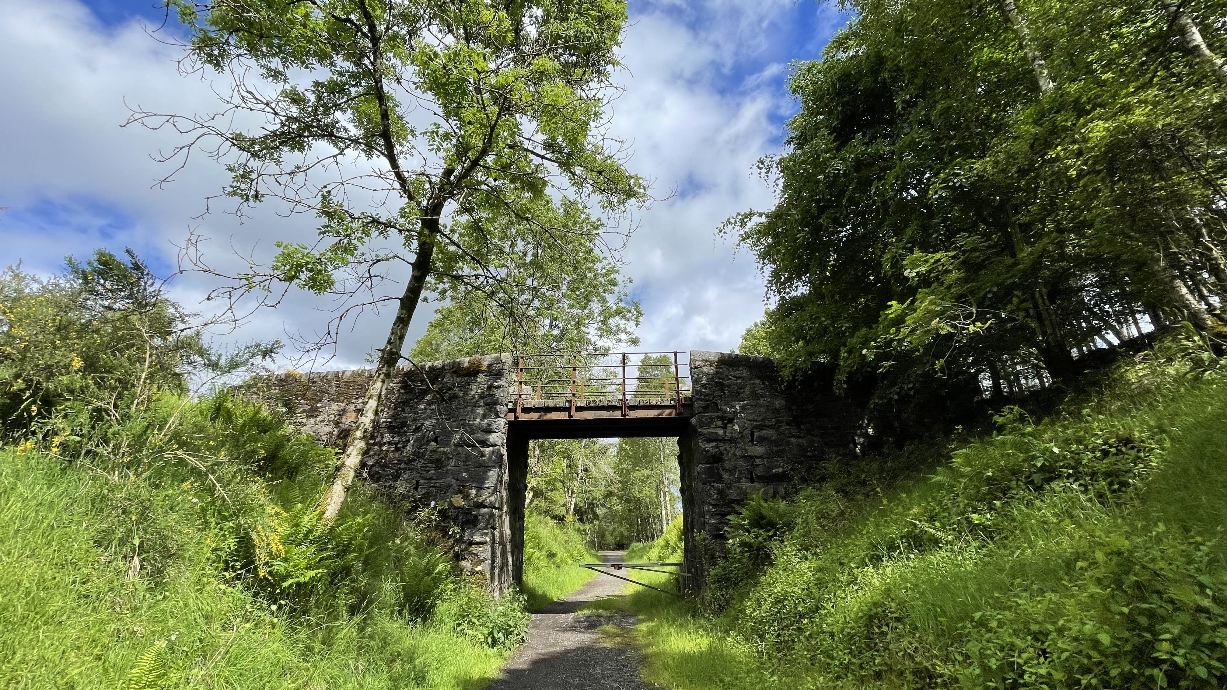 A stone bridge with an iron rail crossing over a lush green trail surrounded by thick trees and dappled sunlight.