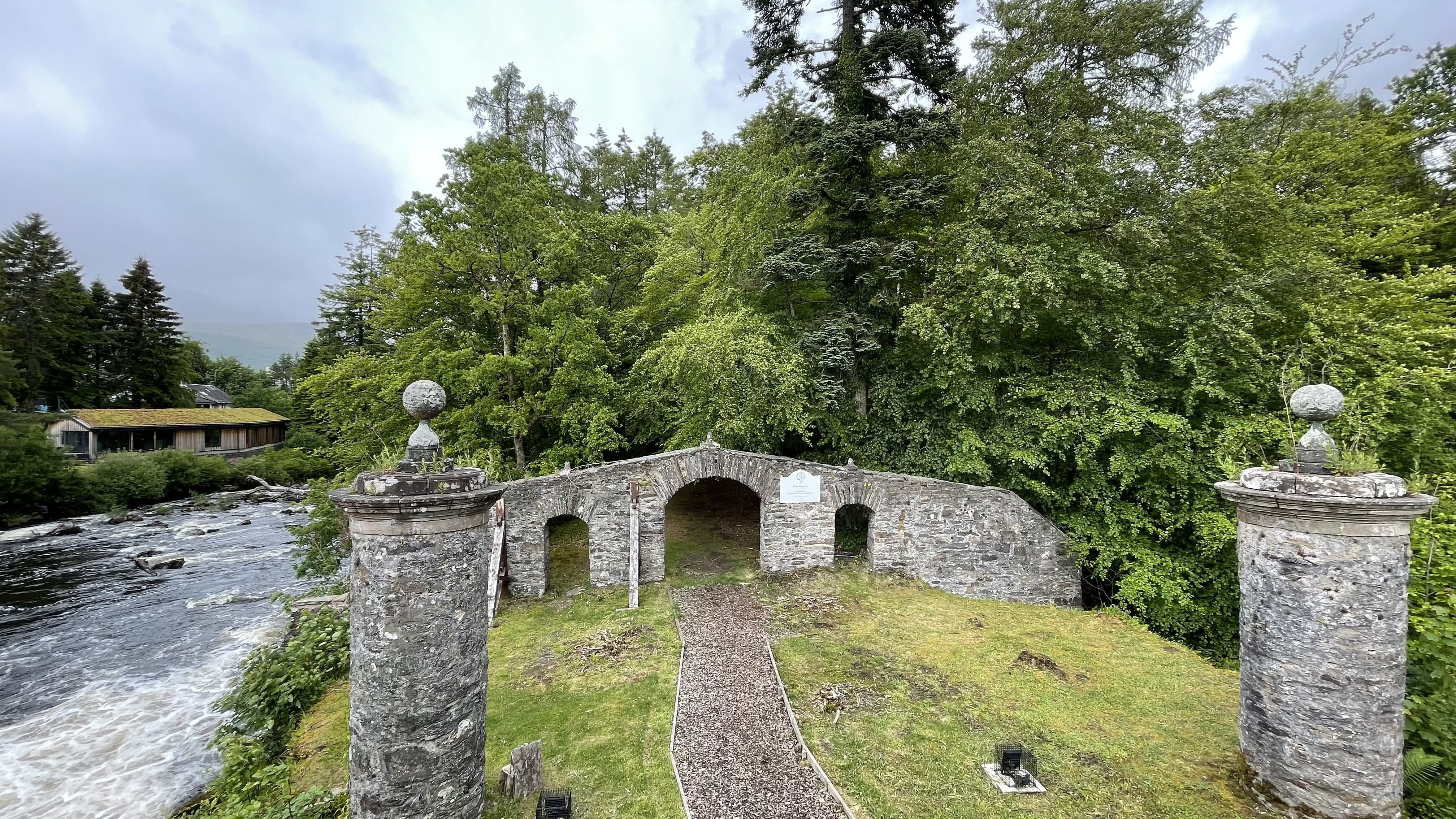 Old stone bridge structure with arches and decorative stone columns, set beside a river and surrounded by trees.