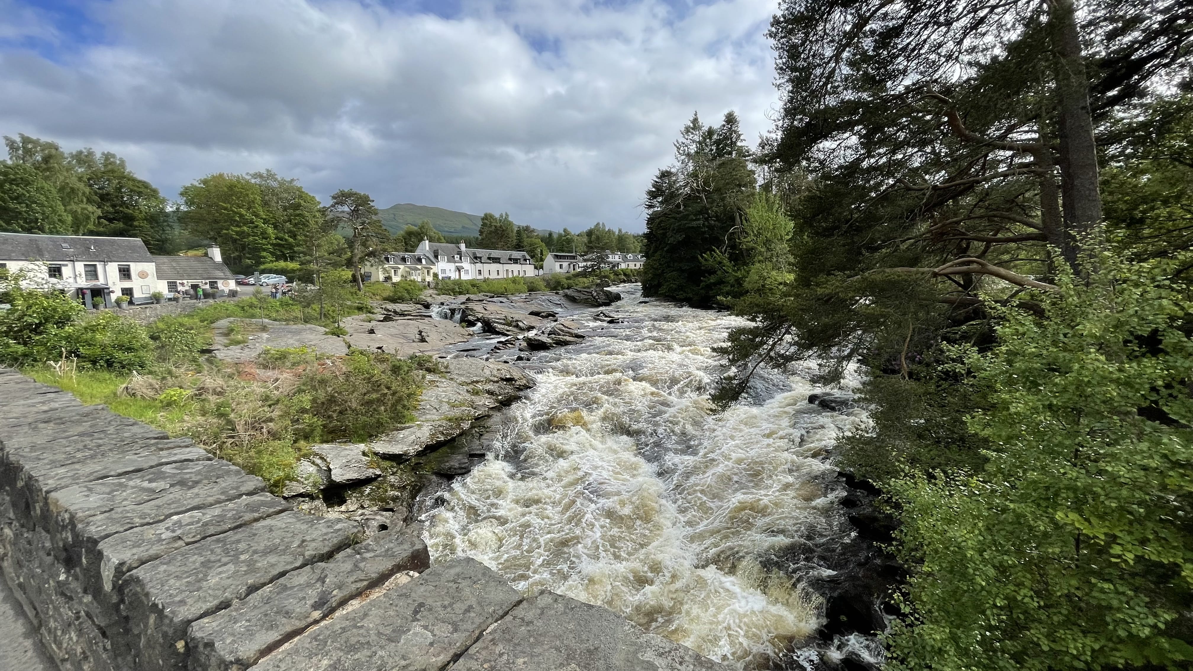 Churning rapids of the River Dochart flowing past whitewashed stone buildings and cottages in Killin, Scotland, viewed from a stone bridge under a partly cloudy sky.