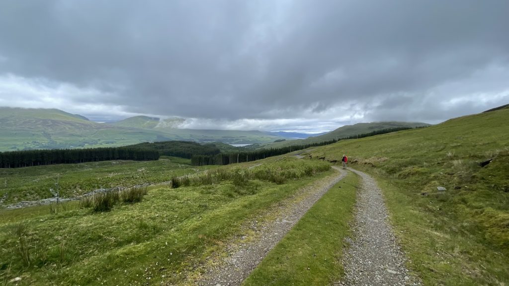 A lone hiker walks along a gravel trail winding through the green hills of the Scottish Highlands under a moody, overcast sky.