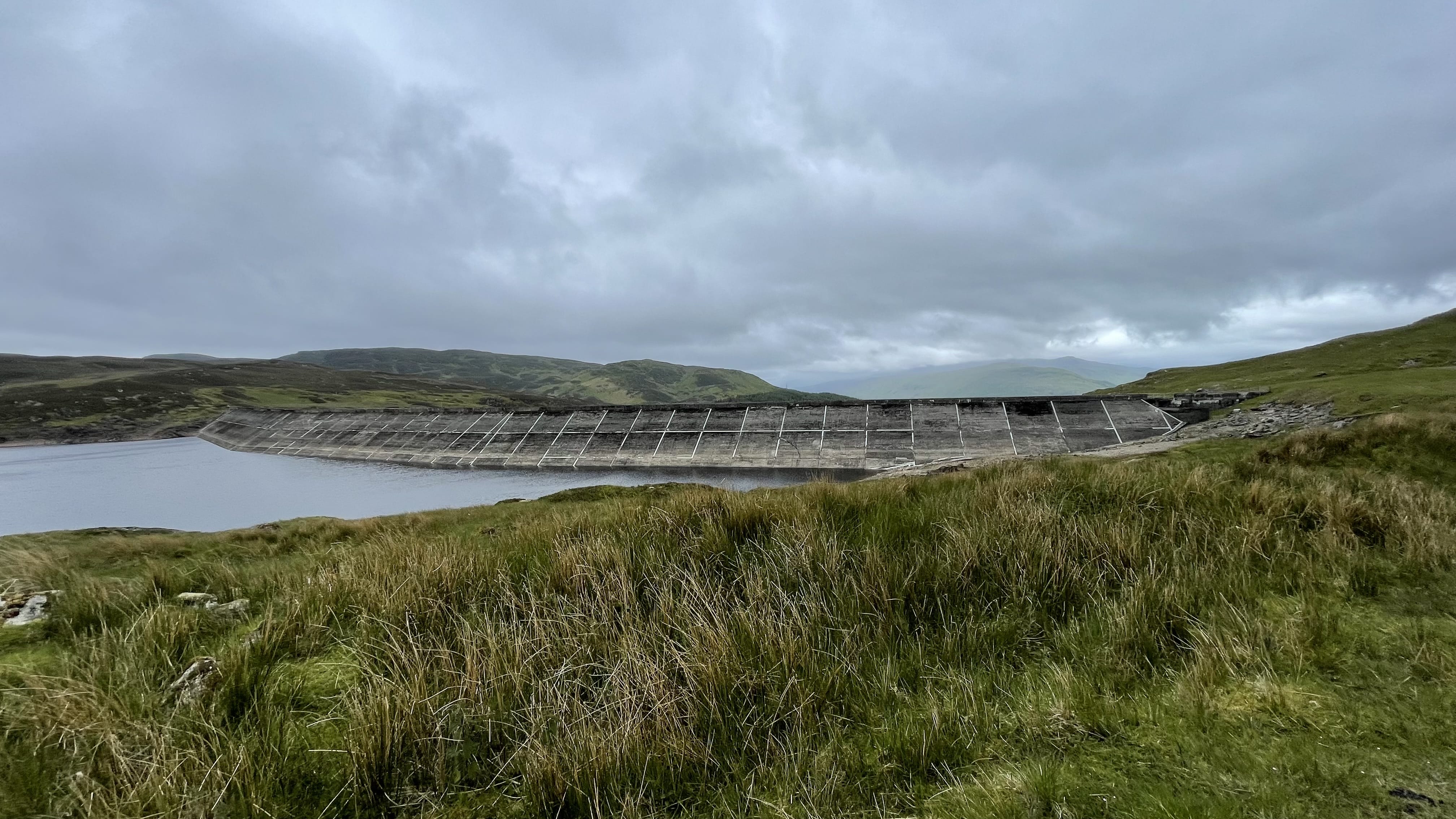 Concrete dam structure at the edge of a remote Scottish loch, surrounded by rolling green hills under a moody, overcast sky.