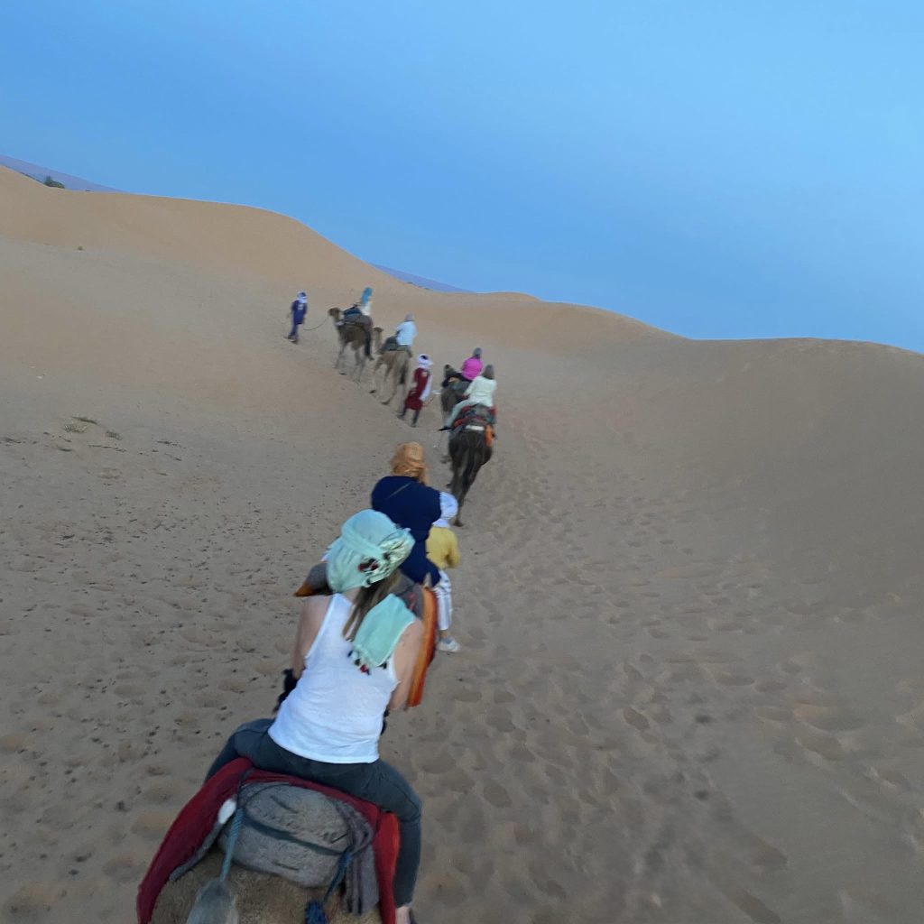 Group of travelers on camels ride single file through soft golden dunes under a pale blue evening sky.