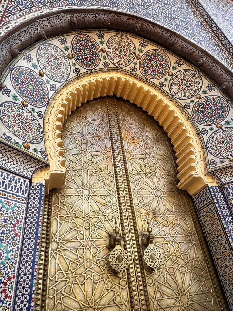 Close-up of a golden brass door at the Royal Palace in Fes, Morocco, featuring intricate geometric patterns, decorative knockers, and colorful mosaic tilework surrounding the arch.