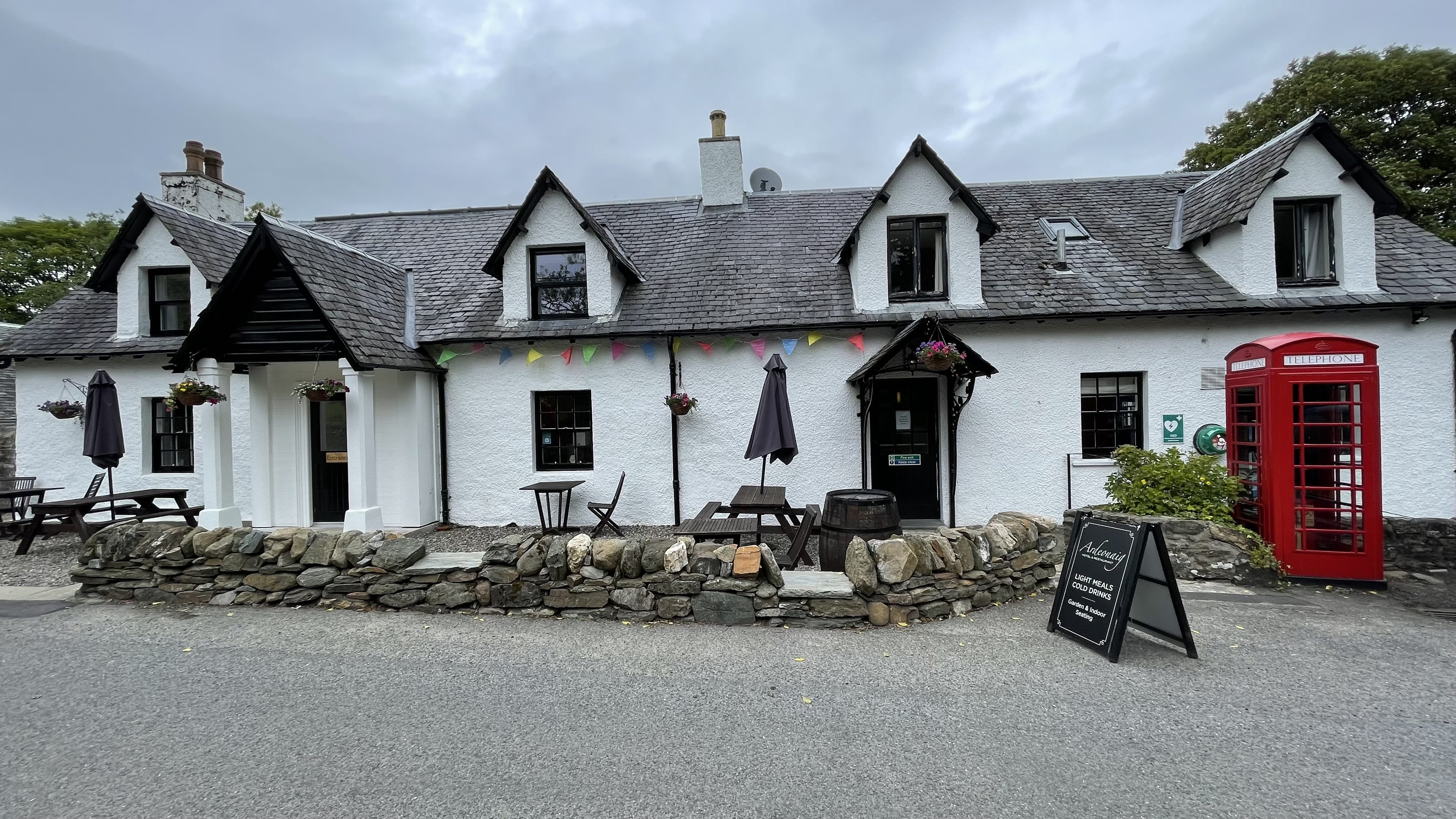 Front view of the Ardeonaig Hotel in Scotland, a whitewashed stone building with hanging flower baskets, outdoor seating, and a classic red telephone box near the entrance.
