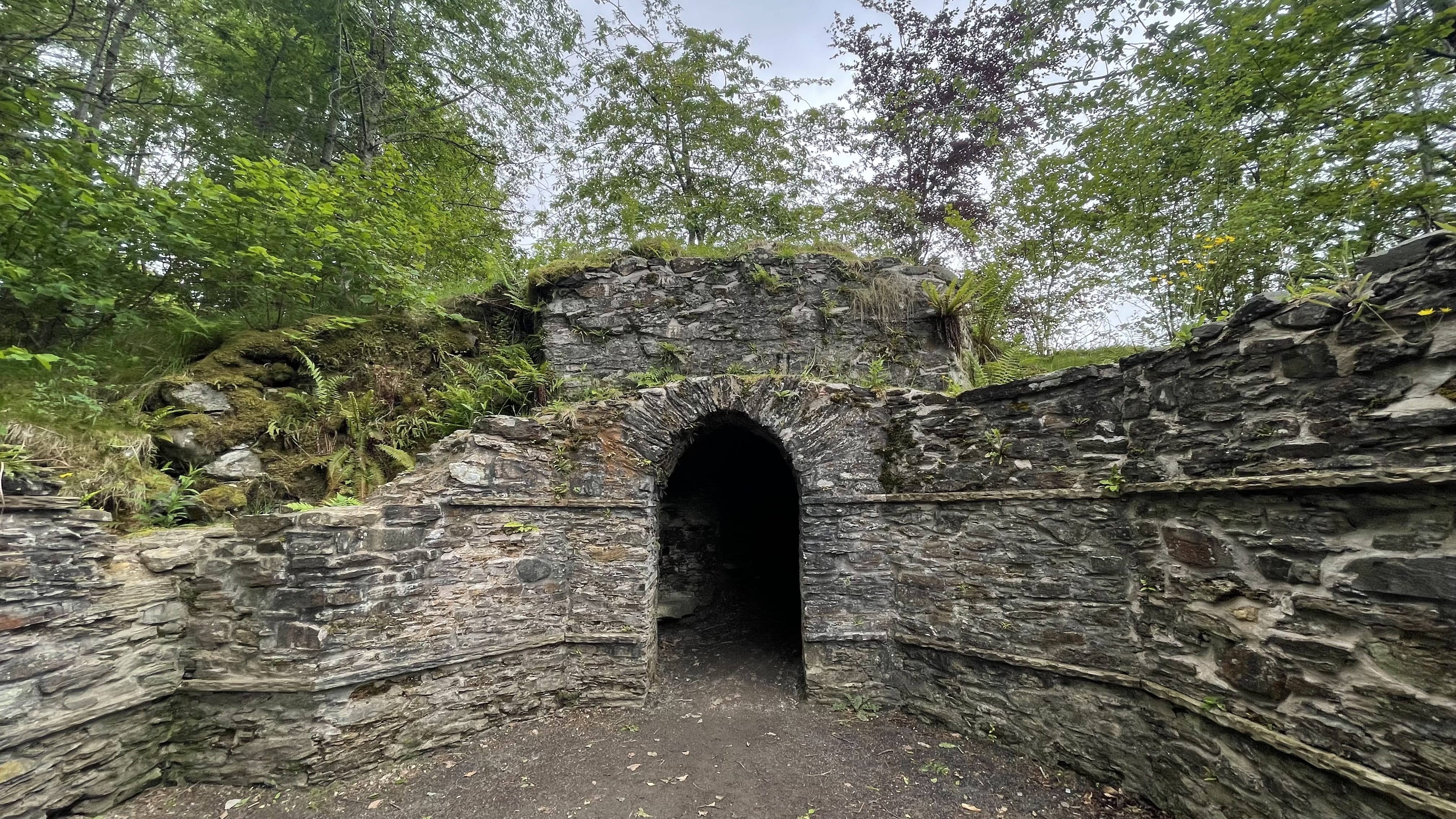 Stone entrance to the Hermit Cave along the Rob Roy Way, surrounded by moss-covered rocks and greenery.
