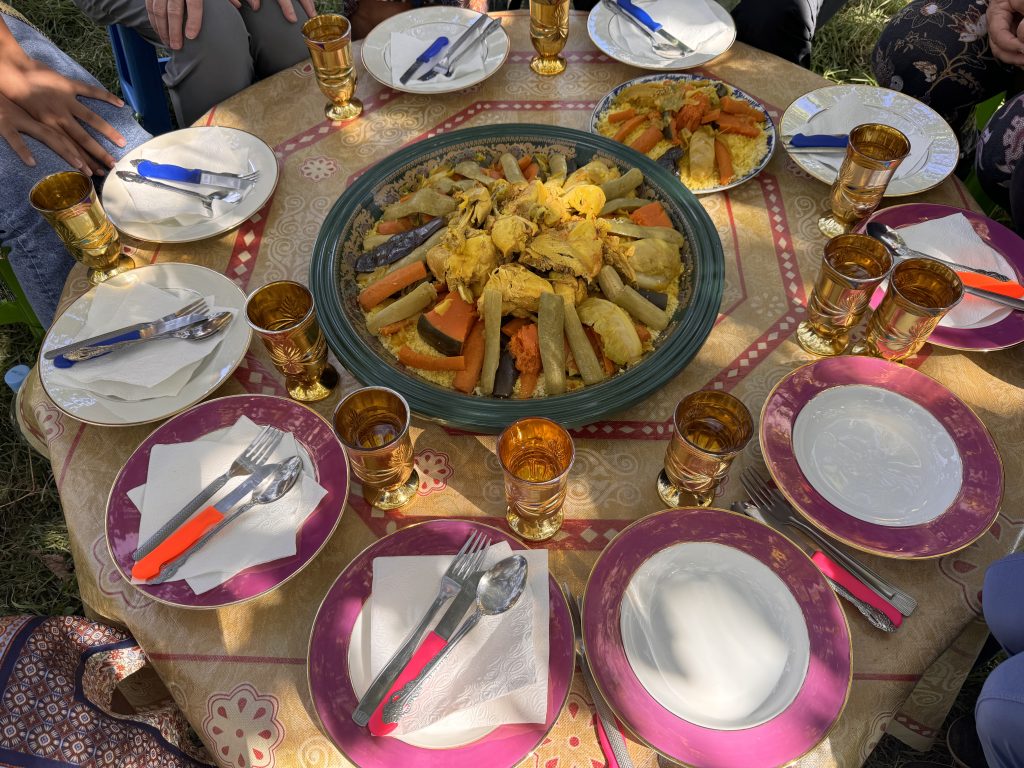 A large communal platter of couscous with vegetables and chicken, surrounded by gold tea glasses and colorful plates in an apple orchard.