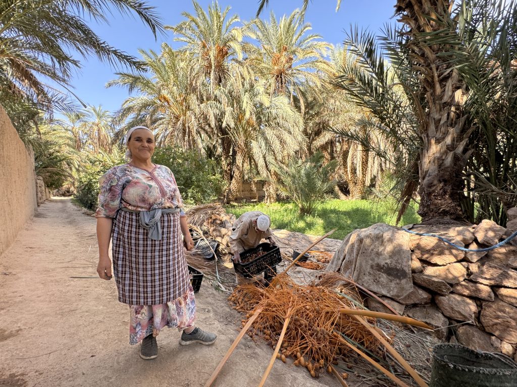 Woman smiling in a dry palm grove with blackened palm trunks and golden hills behind her in the Ziz Valley.