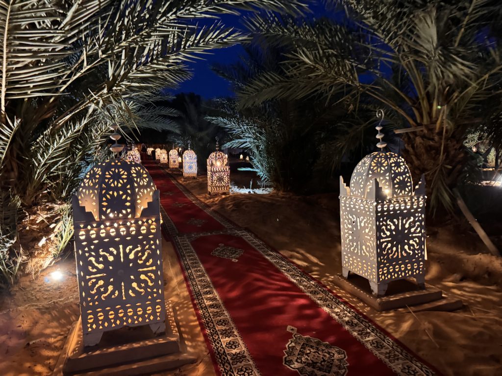 Moroccan lanterns glow warmly along a red carpet leading through a desert camp courtyard under the night sky.