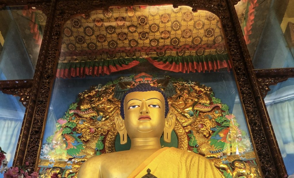 Large golden statue of the Buddha with an intricately decorated backdrop inside a Tibetan temple. Caption: A radiant gold