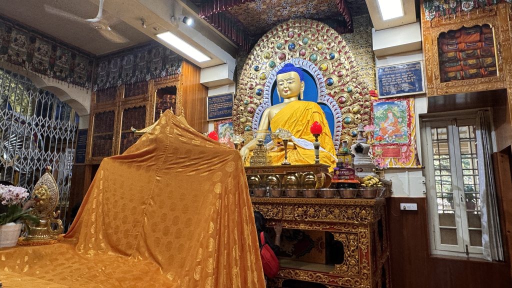 A richly decorated Buddha statue draped in golden robes sits in a colorful shrine adorned with offerings and sacred texts at a Tibetan monastery in McLeod Ganj.