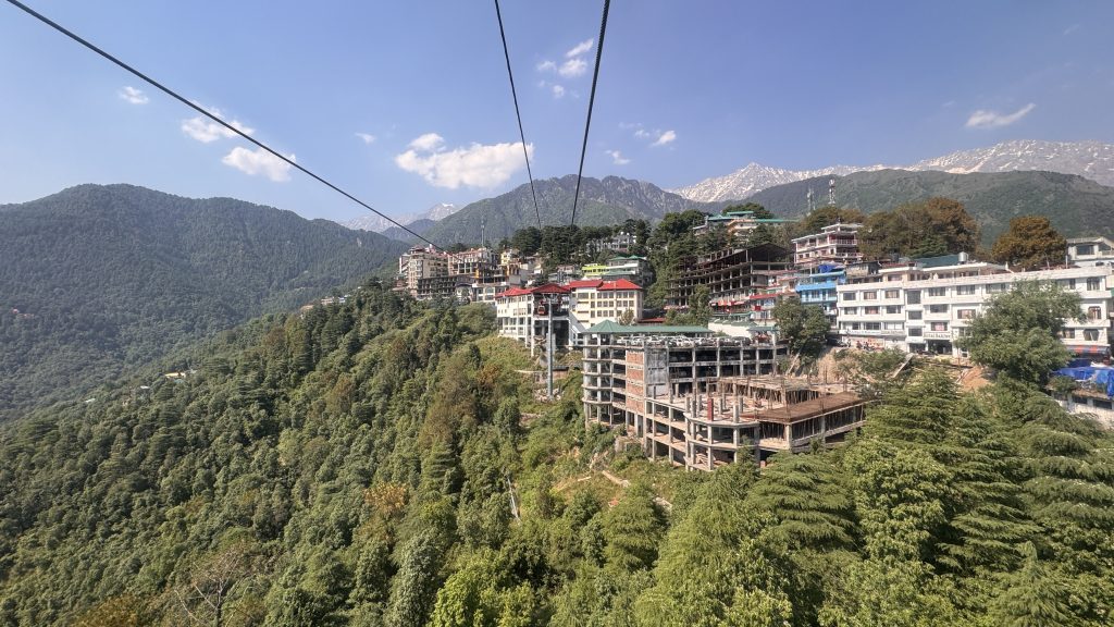 A panoramic view of buildings along the ridge in McLeod Ganj, taken from a cable car with forested hills and mountains in the background