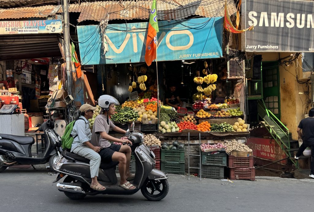 A busy street market in Lower Dharamsala with a colorful fruit stand and two people riding a scooter in the foreground
