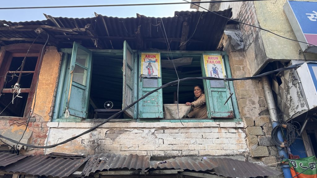 A woman working at a sewing machine sits by open turquoise windows in an old brick building labeled “Arun Tailor”