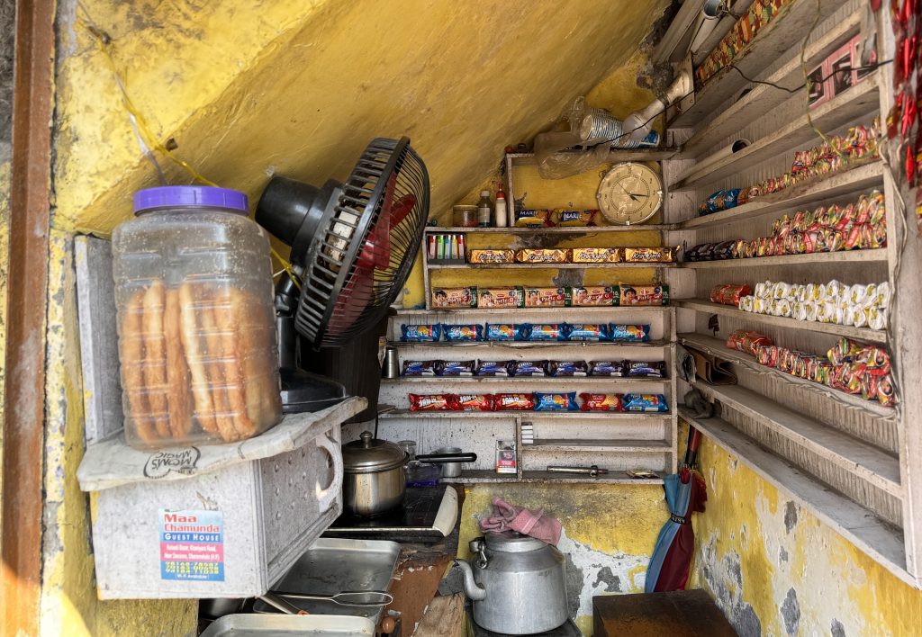 A tiny street-side snack shop with neatly organized biscuit packs and a metal kettle under a yellow ceiling