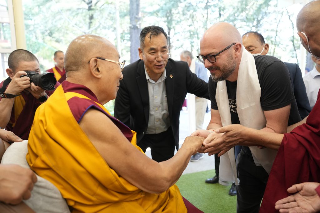 The Dalai Lama holds the hand of a visitor wearing a white ceremonial scarf, surrounded by monks and aides.
