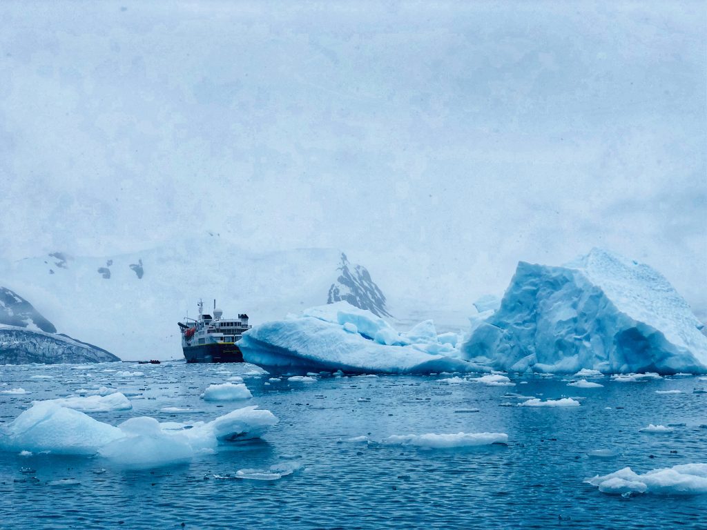 The National Geographic Explorer ship navigating through icy Antarctic waters, surrounded by large icebergs and snow-covered mountains.