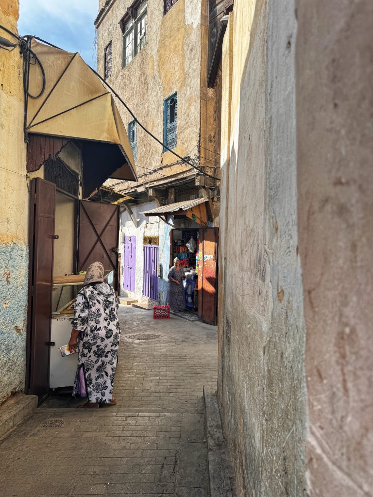 Two women in traditional clothing walking through a narrow alleyway in the Fes medina, surrounded by weathered walls and shops, captured during 2 Days in Fes.