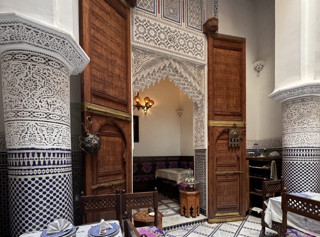 Interior view of Darori Resto in Fes, Morocco, showcasing a traditional Moroccan dining space with intricate zellij tilework, carved plaster columns, arched wooden doors, and a table set with blue and white patterned plates and folded napkins.