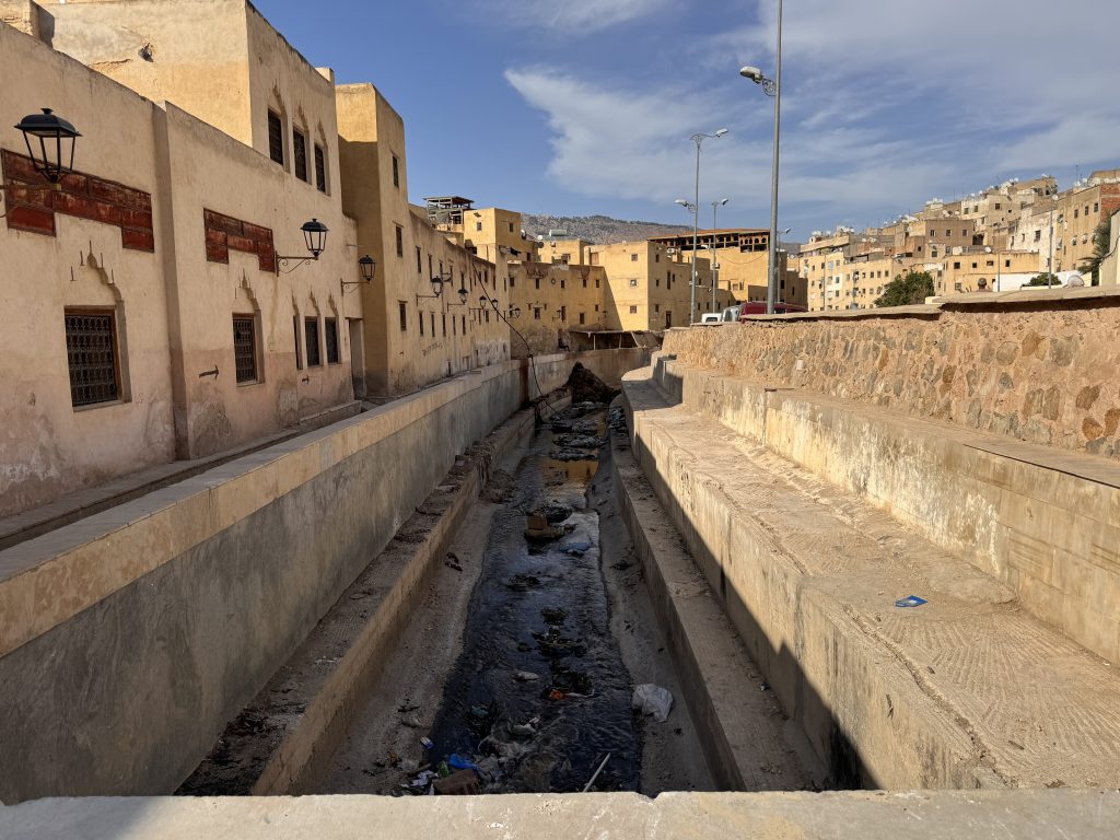 Narrow concrete canal lined with tan buildings in Fes, Morocco, viewed during 2 Days in Fes.