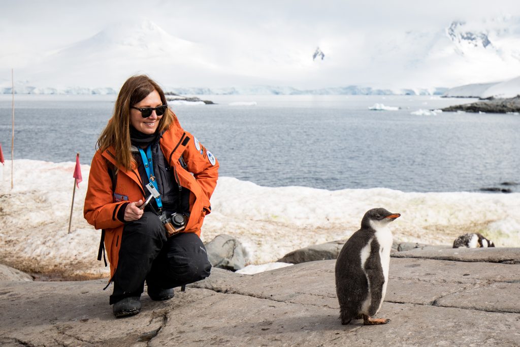 Woman in an orange parka crouching next to a fluffy penguin chick on a rocky shore during an Antarctica cruise, with snow, ocean, and distant mountains in the background.