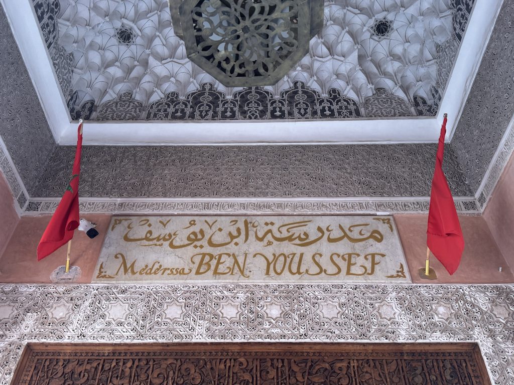 Ornate entrance sign for Medersa Ben Youssef in Marrakech, with Arabic calligraphy and Moroccan flags.