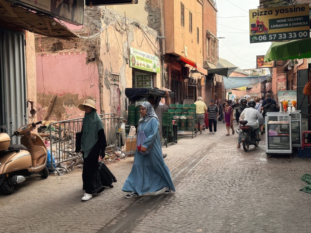 Busy street scene in Marrakech’s medina, with locals and tourists walking past scooters, food stalls, and shop signs – 2 Days in Marrakech.
