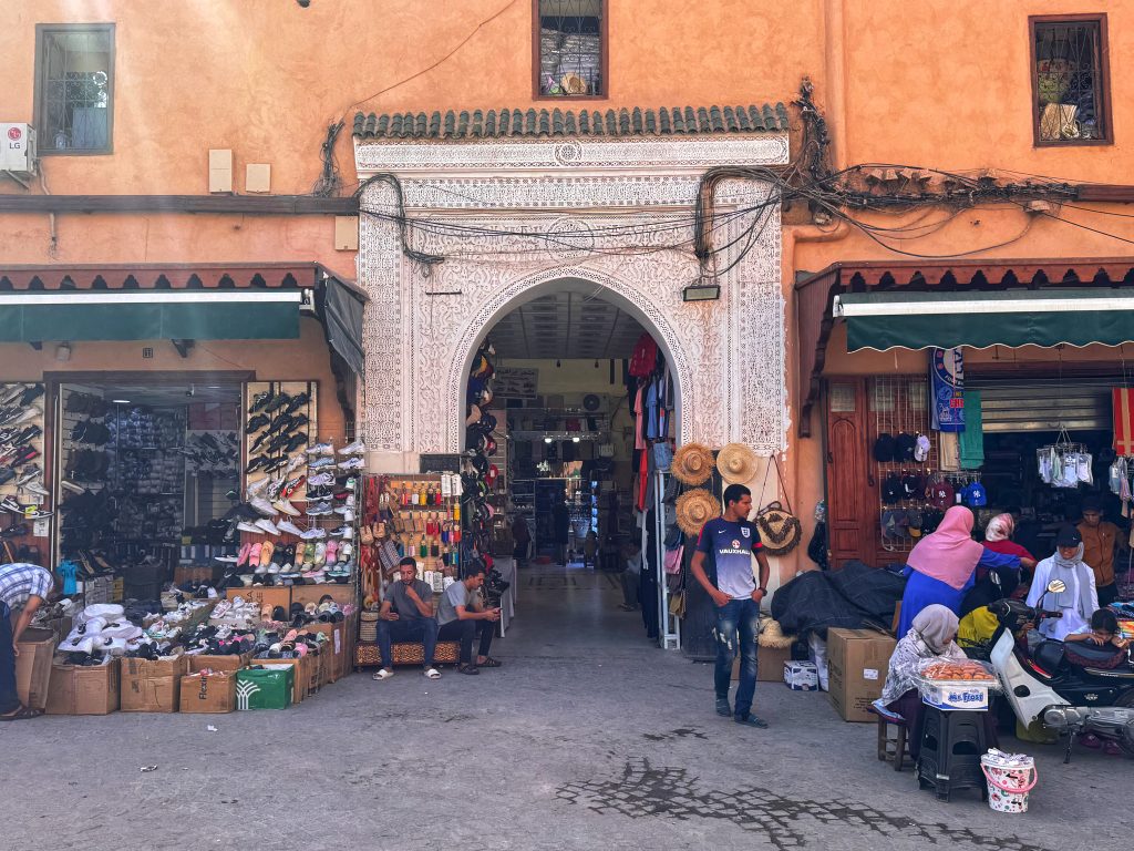 A decorative white archway leading into a Marrakech souk, surrounded by shoe and hat stalls.