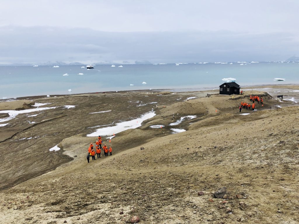 Small groups of people in bright orange parkas walking toward a black wooden hut on Snow Hill Island, Antarctica, with icebergs in the distance and scattered patches of snow on the barren ground.
