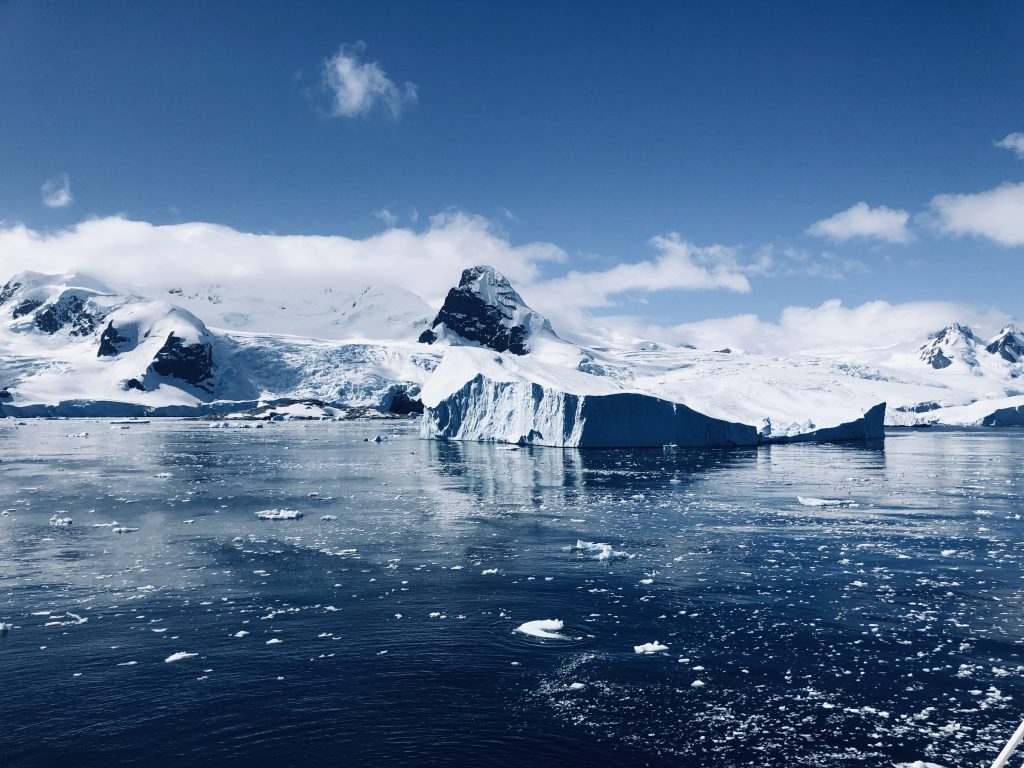 Snow-covered Antarctic mountains and glaciers reflected in deep blue water, with icebergs and scattered ice floating on the surface under a partly cloudy sky.